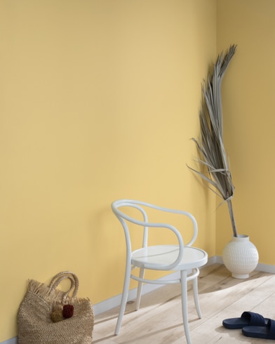 A palm frond, straw tote bag, black slide sandals and white metal chair lean against a wall painted Westminster Gold.