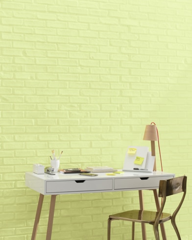 A white and copper writing desk and a bronze metal chair sit in front of a brick wall painted Wales Green.