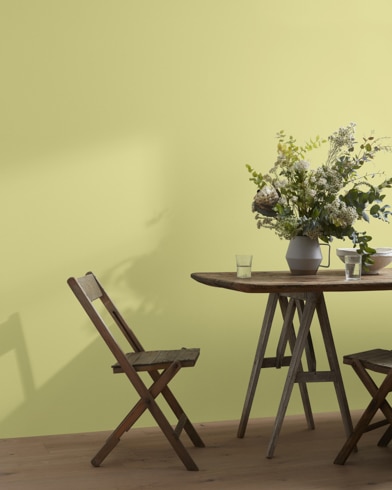 A wood folding chair sits next to a table topped with a greenery-filled vase in front of a Pale Avocado-painted wall.