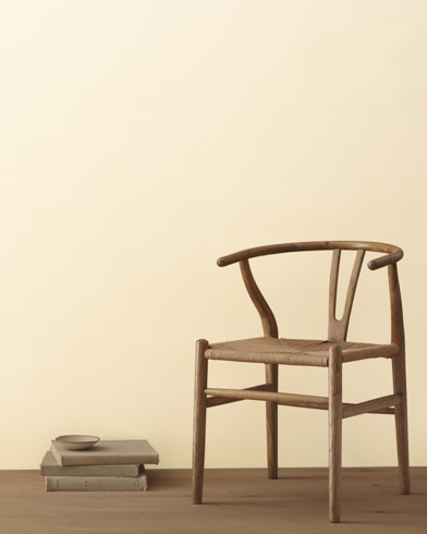 A modern wicker chair and a stack of books topped with a small bowl sit in front of a room painted Mannequin Cream.