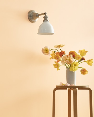 A Golden Dunes-painted wall with single sconce, and a wooden stool with flower-filled vase.