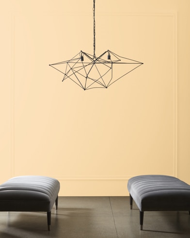 A modern, wiry chandelier hangs over two velvet ottomans in front of a wall painted Wheatfield.