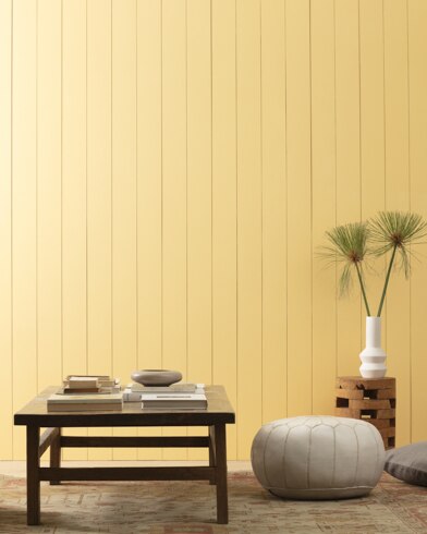 Plush ottomans, a low accent table and a vase with wispy greens sit in front of a paneled wall painted Governor's Gold.