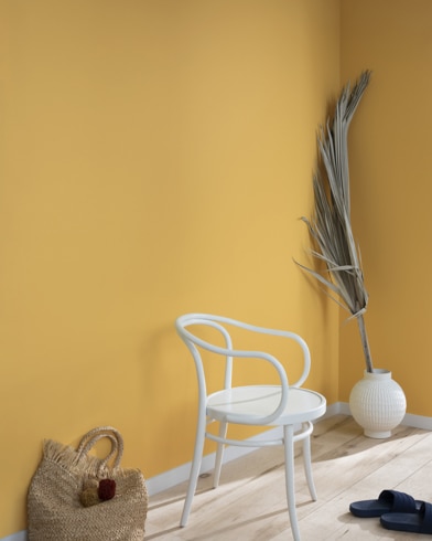 A palm frond, straw tote bag, black slide sandals and white metal chair lean against a wall painted Stuart Gold.