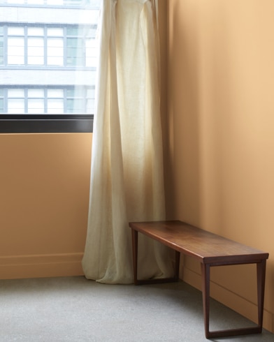 In a Freenfield Pumpkin-painted room with a wooden bench, a gauzy white curtain hangs next to an open window.