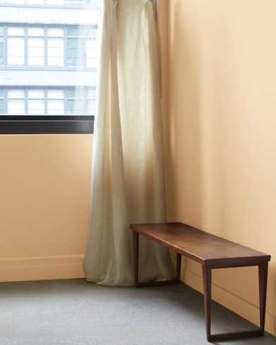 In a Roxbury Caramel-painted room with a wooden bench, a gauzy white curtain hangs next to an open window.