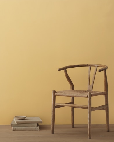A modern wicker chair and a stack of books topped with a small bowl sit in front of a room painted Chestertown Buff.